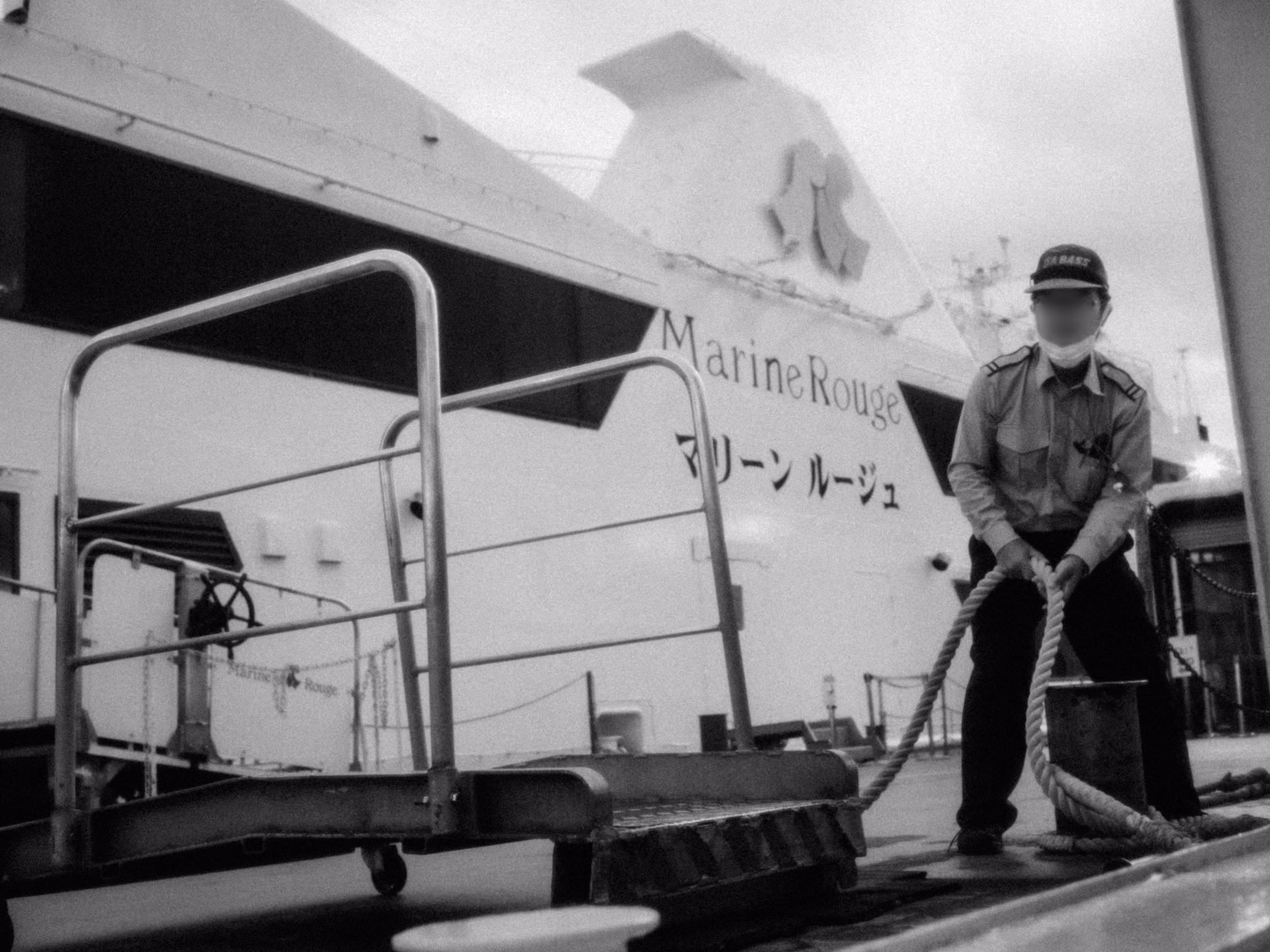 A masked Marine Rouge deckhand bracing with both hands to pull a thick mooring rope, the cruise ship behind him.