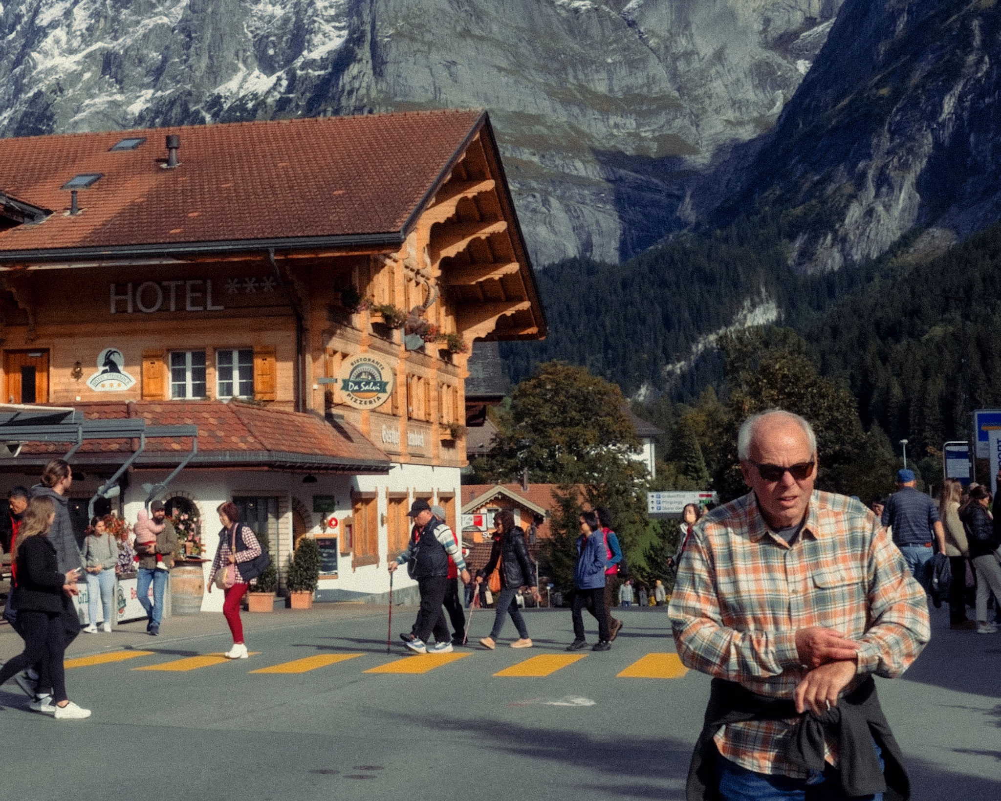 Elderly man in checkered shirt walking through town square with traditional Alpine hotel and dramatic mountain cliff in background.