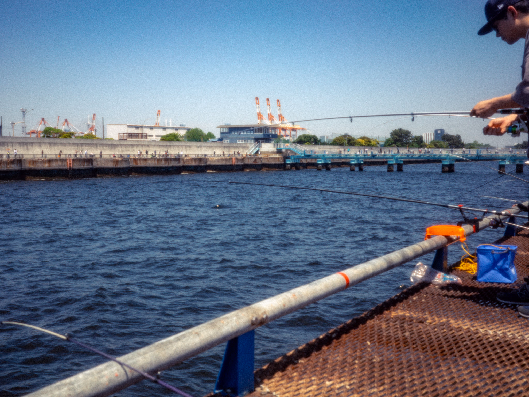 Angler casting a fishing rod into the sea from Honmoku Pier under a clear blue sky, with port cranes across the water.