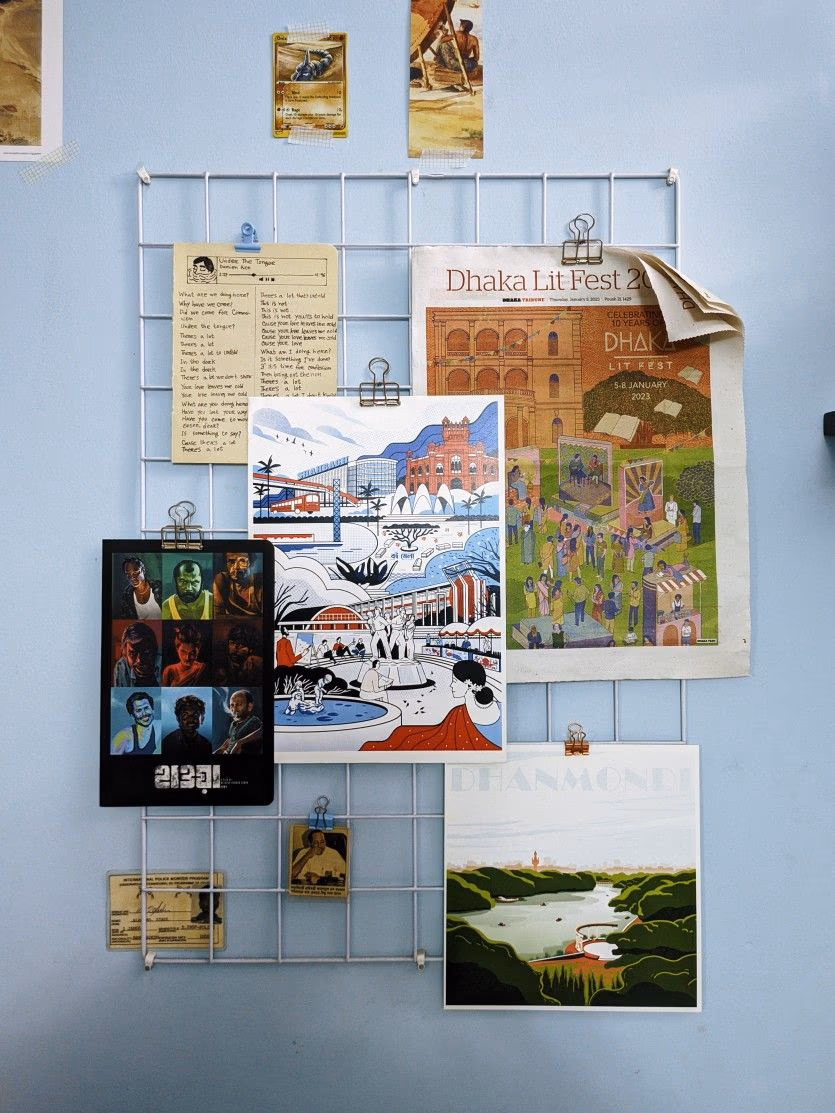 A white wire grid panel mounted on a pale blue wall, hung with bulldog-clipped print: a handwritten note on yellow paper, a newspaper page for a Dhaka literature festival, an illustrated city print, a dark grid of film portraits, and a landscape print, with a few small cards taped to the wall above the grid.